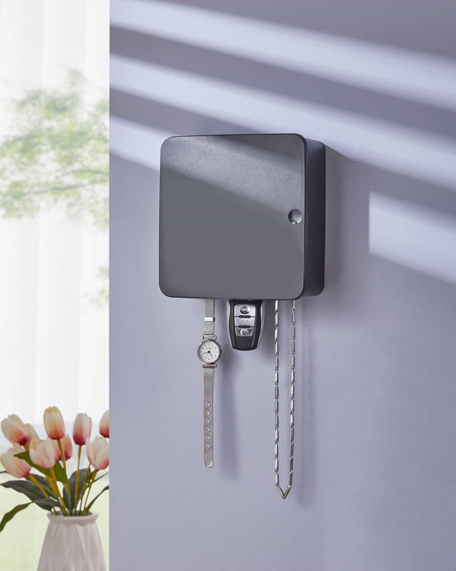 A matte black wall-mounted key organizer box holding a watch, car key, and chain on a pastel wall with tulip flowers in the background.