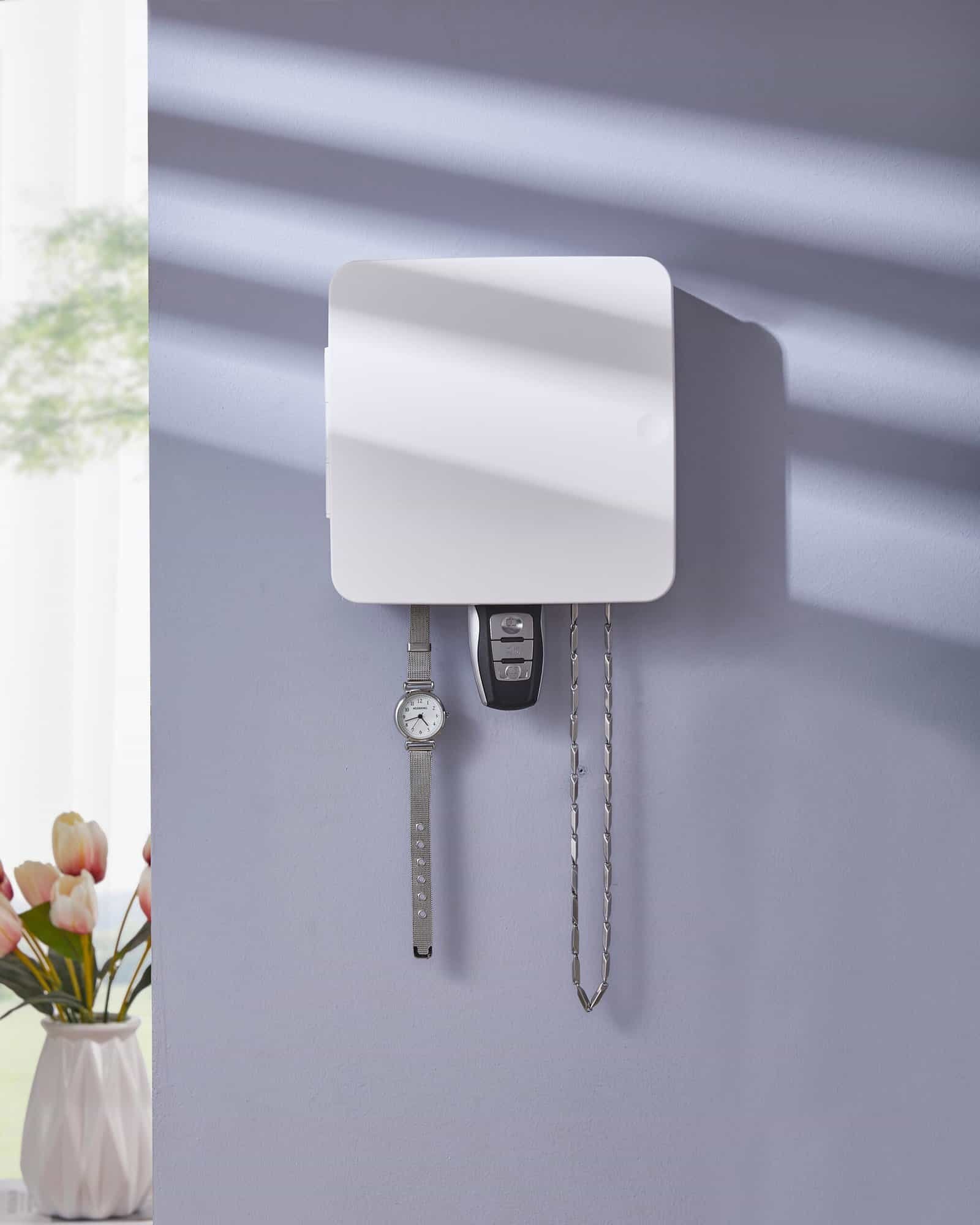 A matte white wall-mounted key organizer box holding a watch, car key, and chain on a pastel wall with tulip flowers in the background.