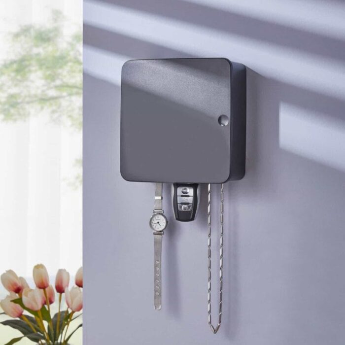 A matte black wall-mounted key organizer box holding a watch, car key, and chain on a pastel wall with tulip flowers in the background.